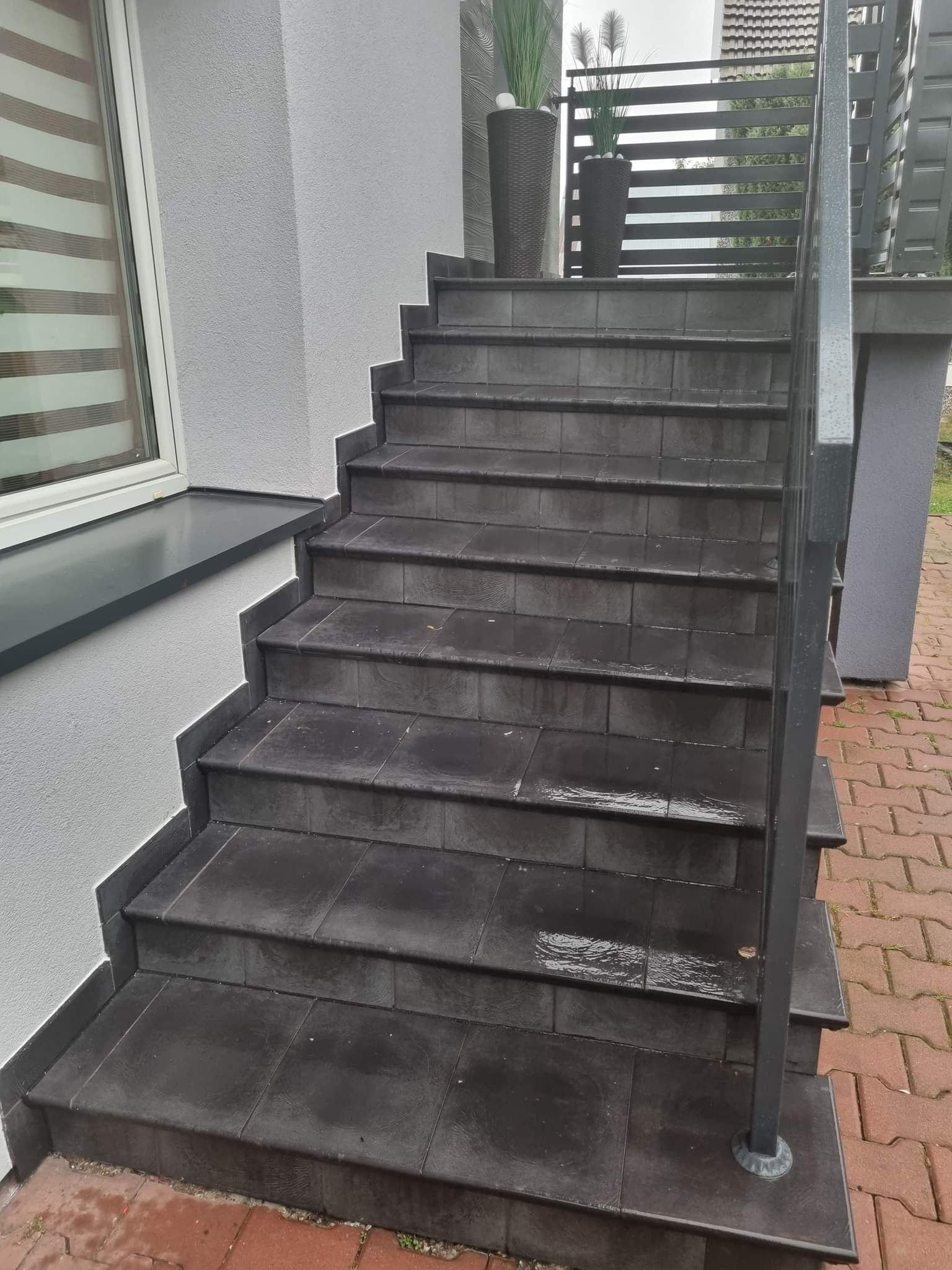 Zewnętrzne schody wyłożone ciemnymi płytkami, z metalową barierką, prowadzące do wejścia do domu, z donicami z roślinami po bokach.