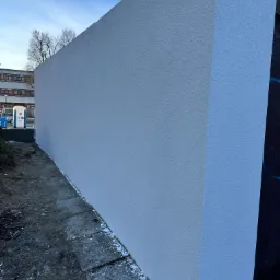 Świeżo nałożona, jasnoszara elewacja z wyraźną fakturą, widoczna z boku, z fragmentem budynku w tle i przenośną toaletą w oddali.