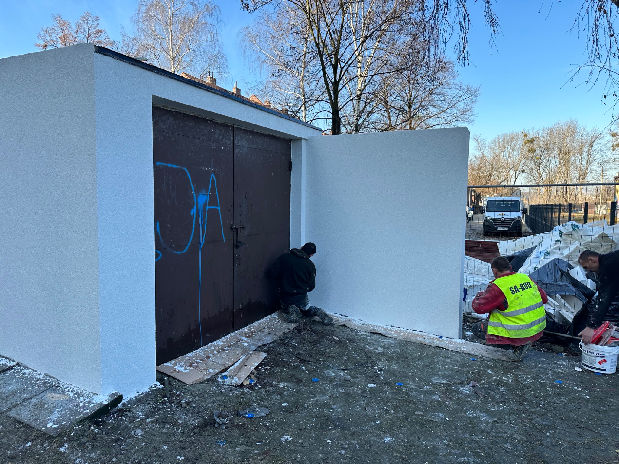 Dwaj pracownicy w trakcie nakładania białej struktury na elewację garażu z brązowymi drzwiami pokrytymi graffiti; jeden z nich w kamizelce odblaskowej z napisem SA-BUD.