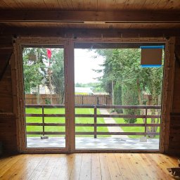 RAJPLAST SYLWIA RAJCHERT - Montaż dwuskrzydłowego okna balkonowego w drewnianym domu w Warszawie. Widok na ogród z drewnianym płotem i trawnikiem.