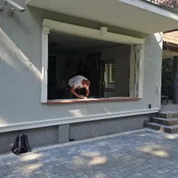 Pracownik przygotowuje otwór okienny do montażu, widoczny fragment elewacji domu z ozdobnym balkonem i kostka brukowa na podjeździe.