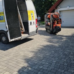 RAJPLAST SYLWIA RAJCHERT - Biały samochód dostawczy z otwartymi drzwiami i naklejką 'Sprzedaż i montaż okien' na tle podjazdu wyłożonego kostką brukową i zaparkowanego mini-dźwigu.