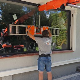 RAJPLAST SYLWIA RAJCHERT - Montaż okna z użyciem pomarańczowego mini dźwigu przez pracownika w szarej koszulce z logo firmy, widok od tyłu na tle domu z ogrodem.