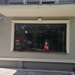 RAJPLAST SYLWIA RAJCHERT - Duże, panoramiczne okno z czarną ramą w elewacji domu; w odbiciu widoczna osoba w czerwonych spodenkach i pomarańczowy podnośnik koszowy.