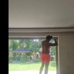 RAJPLAST SYLWIA RAJCHERT - Mężczyzna w krótkich spodenkach, stojąc na parapecie, montuje okno, widok na ogród z altaną.