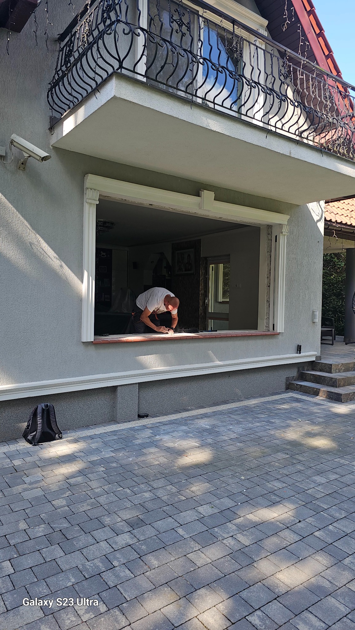 Pracownik przygotowuje otwór okienny do montażu, widoczny fragment elewacji domu z ozdobnym balkonem i kostka brukowa na podjeździe.