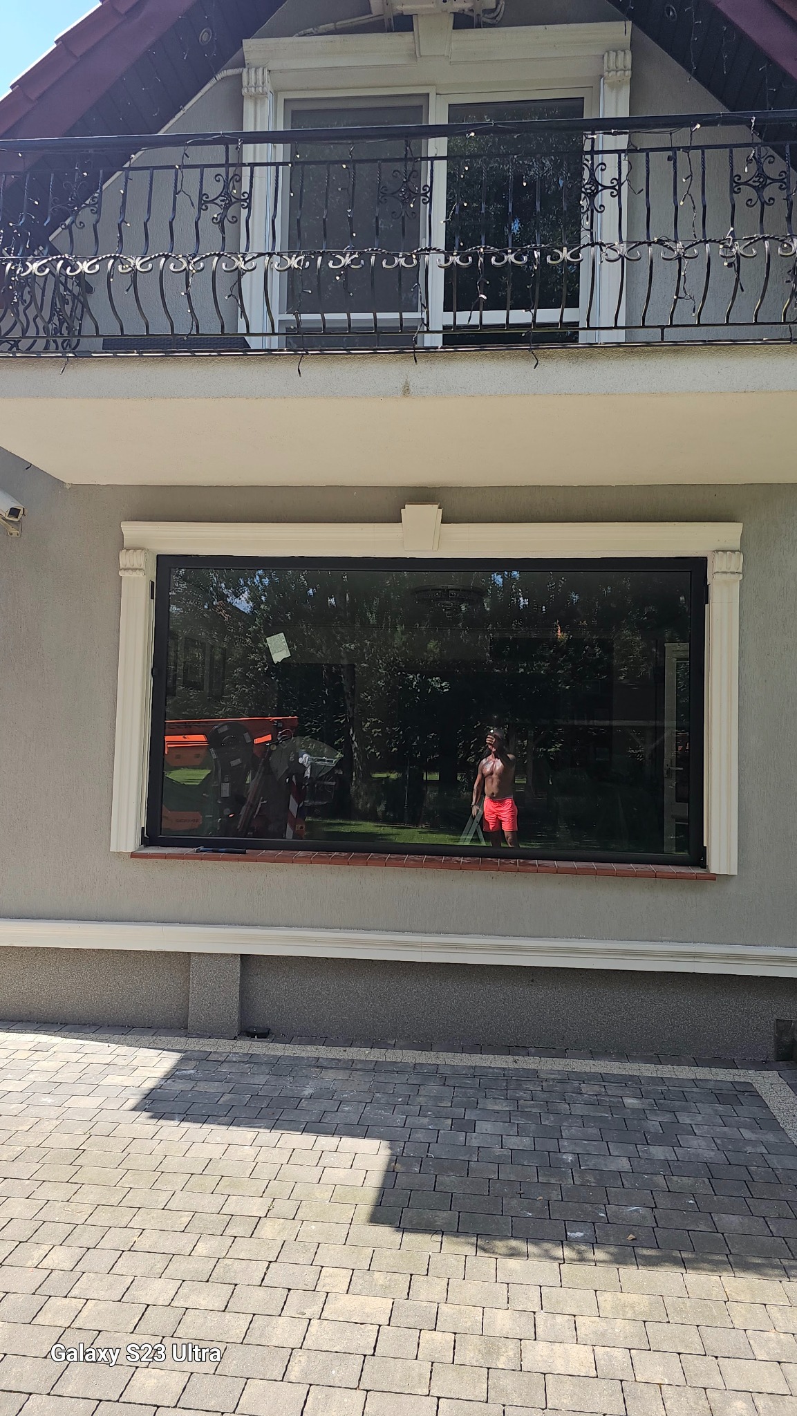 Duże, panoramiczne okno z czarną ramą w elewacji domu; w odbiciu widoczna osoba w czerwonych spodenkach i pomarańczowy podnośnik koszowy.