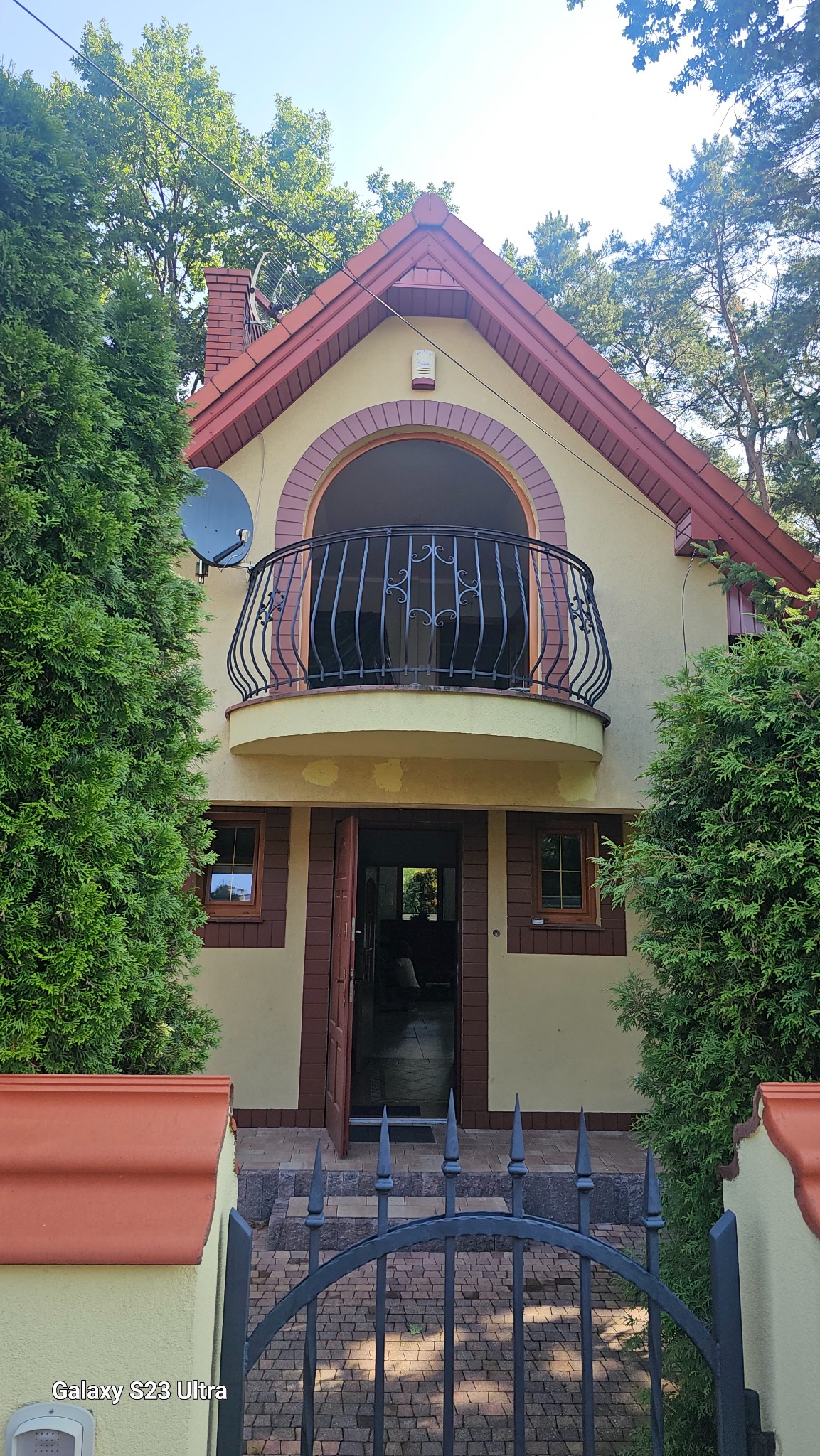 Dwupiętrowy dom z balkonem z kutą balustradą, widok zza metalowego ogrodzenia, otoczony zielenią.