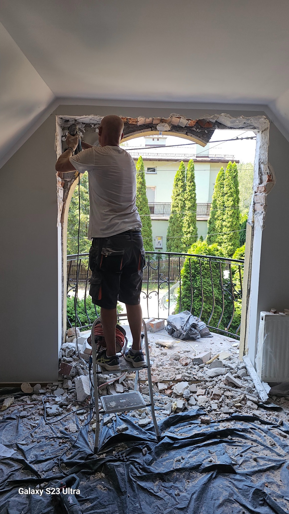 Mężczyzna na drabinie, w trakcie demontażu starego okna z łukiem, widok z wnętrza, gruz zabezpieczony folią, balkon z ozdobną balustradą na zewnątrz.