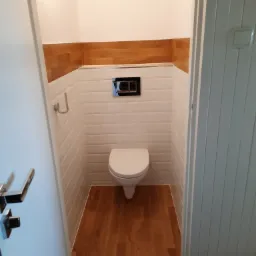 Mała toaleta po remoncie, z drewnianym panelem na górze ściany, białymi płytkami ułożonymi w cegiełkę i drewnopodobną podłogą, widok zza otwartych drzwi.