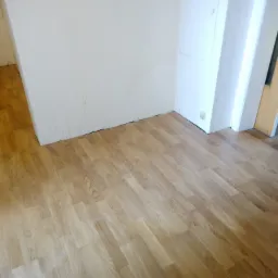 Jasne panele podłogowe ułożone w pokoju z białymi ścianami i widocznym fragmentem drzwi.