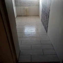 Ułożone jasne, podłużne płytki imitujące drewno na podłodze w łazience, widoczny grzejnik na ścianie z białej cegiełki i fragment dekoracyjnej ściany z wzorem.