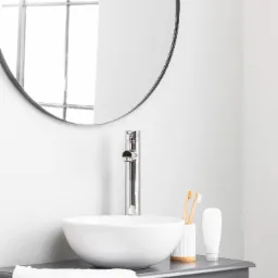 Minimalistyczna łazienka z okrągłym lustrem, białą umywalką nablatową, chromowaną baterią, bambusowymi szczoteczkami do zębów i szarą szafką pod umywalką.