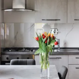 Jasnoszara kuchnia z marmurowym backsplash, bukiet tulipanów w szklanym wazonie na stole, obok leży smartfon w czerwonym etui.