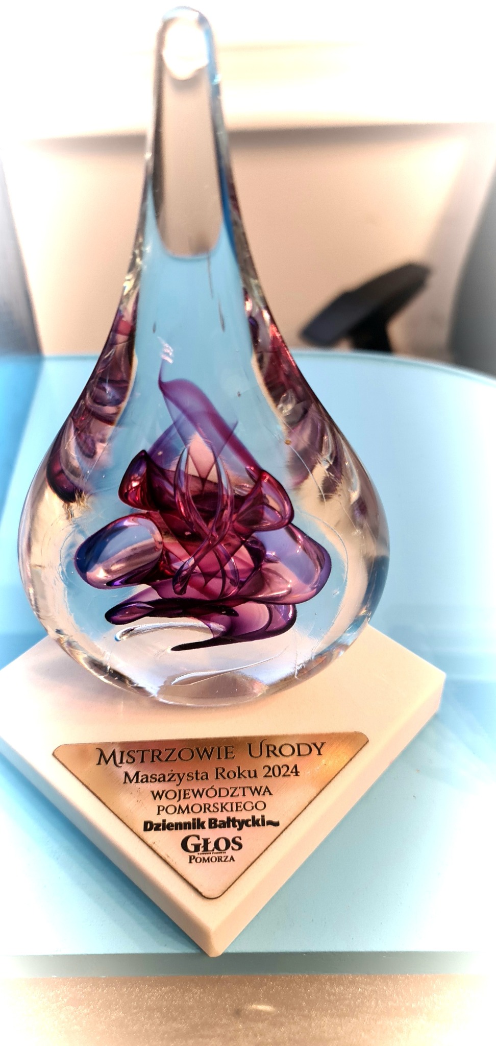 Szklana statuetka w kształcie łzy z fioletową abstrakcyjną formą wewnątrz, na białej podstawie z brązową tabliczką 'Mistrzowie Urody Masażysta Roku 2024 Województwa Pomorskiego'
