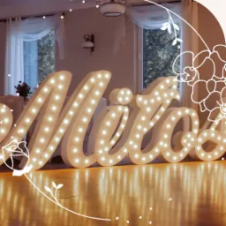 Podświetlany napis 'Miłość' w stylu retro z żarówkami, ustawiony na parkiecie w pomieszczeniu z oknami w tle.