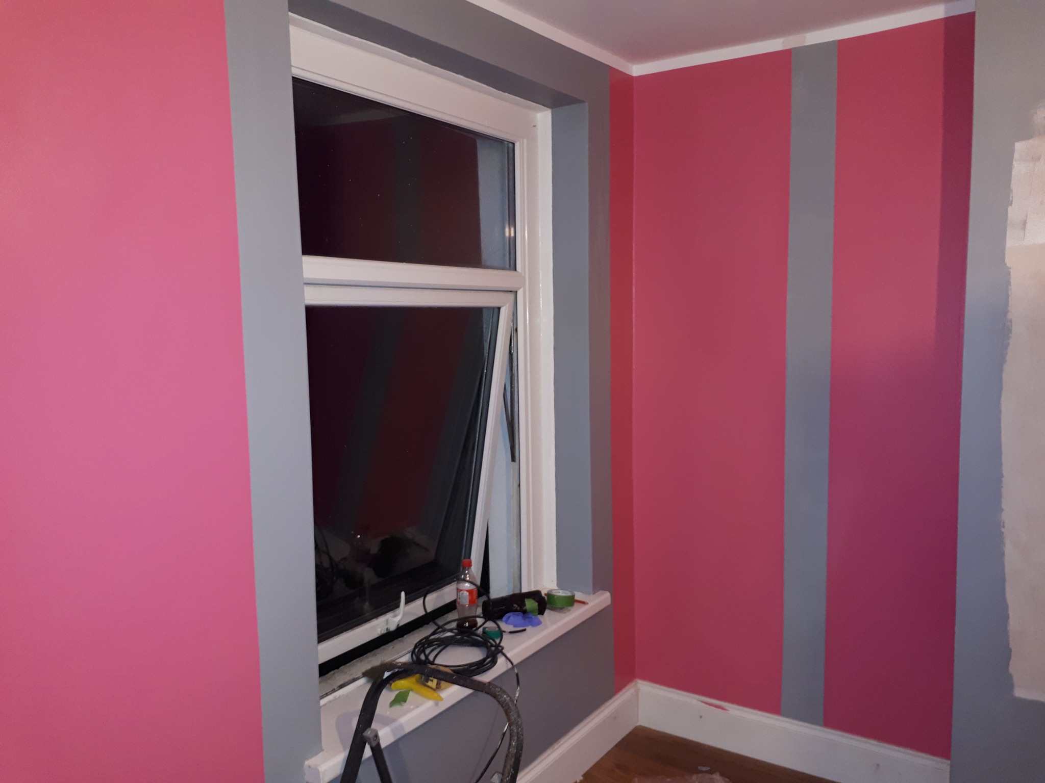 Pokój w trakcie malowania, ściany w odcieniach różu i szarości, widoczne narzędzia malarskie na parapecie okiennym i drabina malarska.