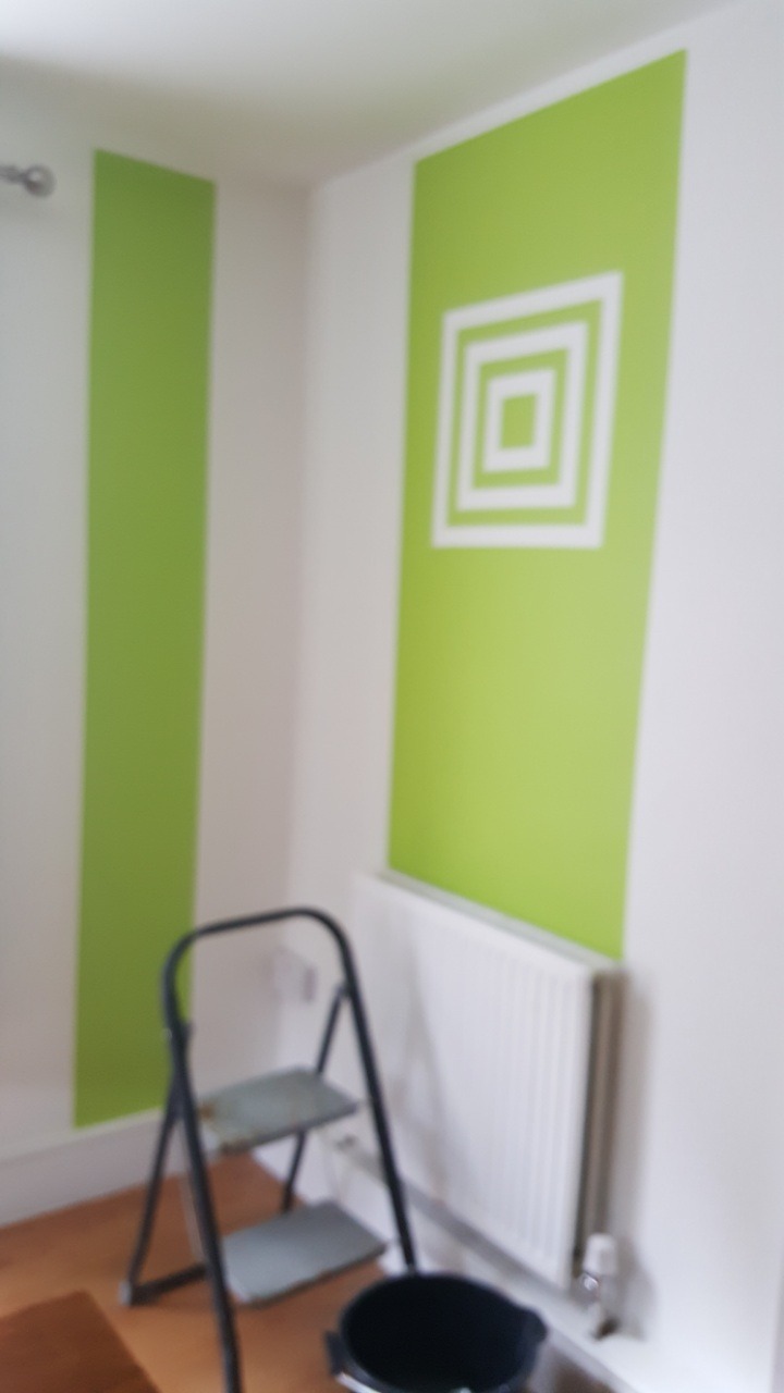 Świeżo pomalowana ściana w pokoju z zielonymi pasami i geometrycznym wzorem w postaci białych kwadratów, drabina malarska i wiadro w rogu pomieszczenia.