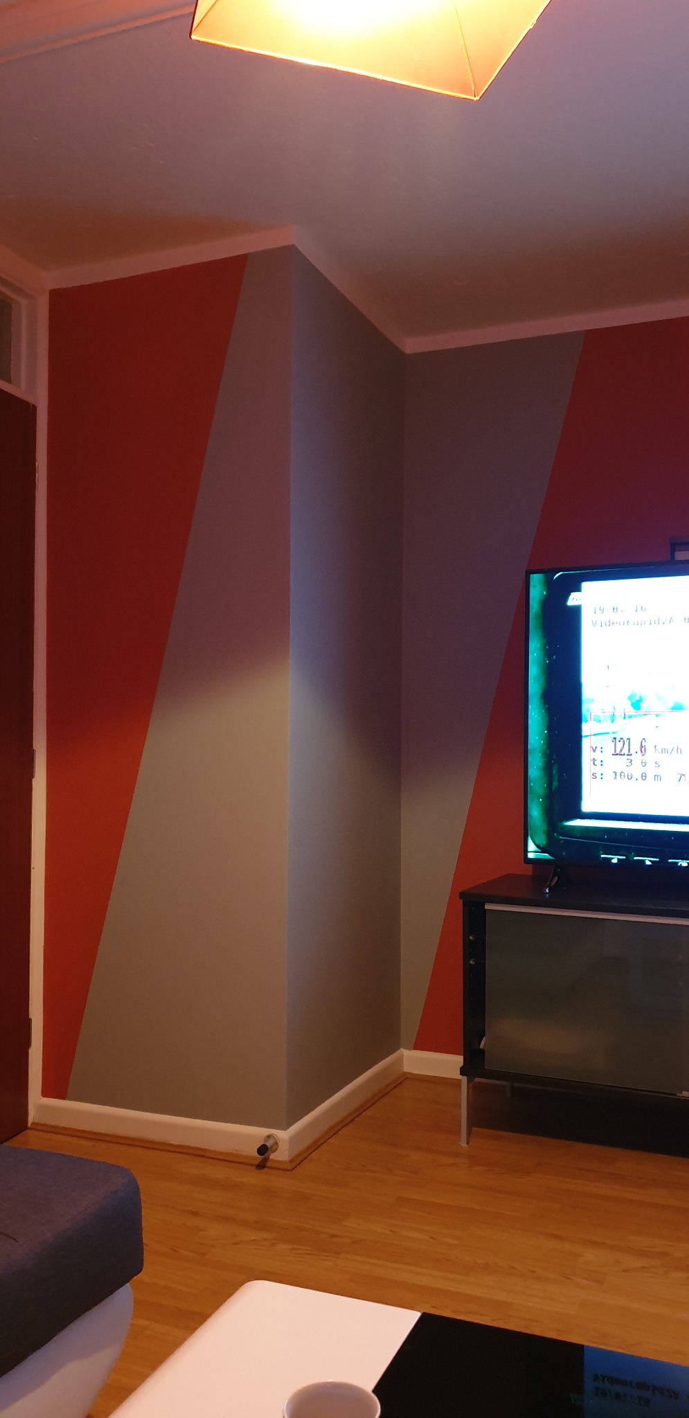 Pomarańczowo-szara ściana z geometrycznym wzorem w rogu pokoju, widoczny fragment telewizora i lampy sufitowej