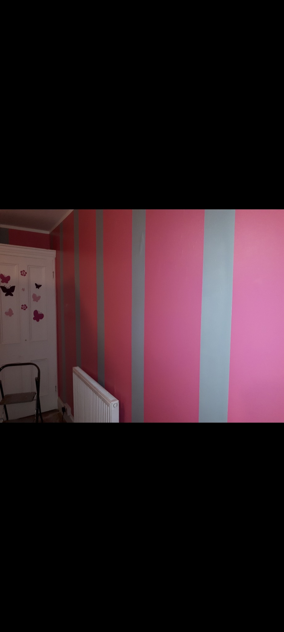 Pokój dziecięcy z pomalowanymi ścianami w szerokie, pionowe pasy koloru różowego i szarego. Przy ścianie stoi biały grzejnik i drabinka malarska, a po lewej stronie widoczne białe drzwi...