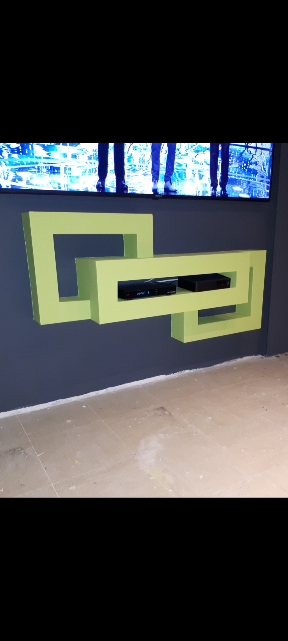 Nietypowa, jasnozielona półka RTV w kształcie połączonych kwadratów, zamontowana na szarej ścianie pod telewizorem. Na półce widoczne dwa czarne dekodery. Na podłodze widoczne ślady po farbie.