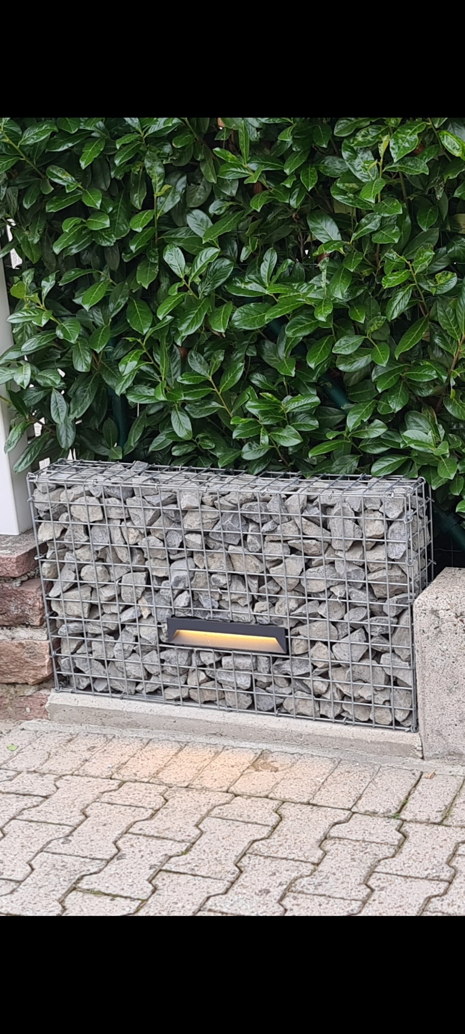 Gabion z szarego kamienia z wbudowanym oświetleniem punktowym na tle zielonego żywopłotu i kostki brukowej.