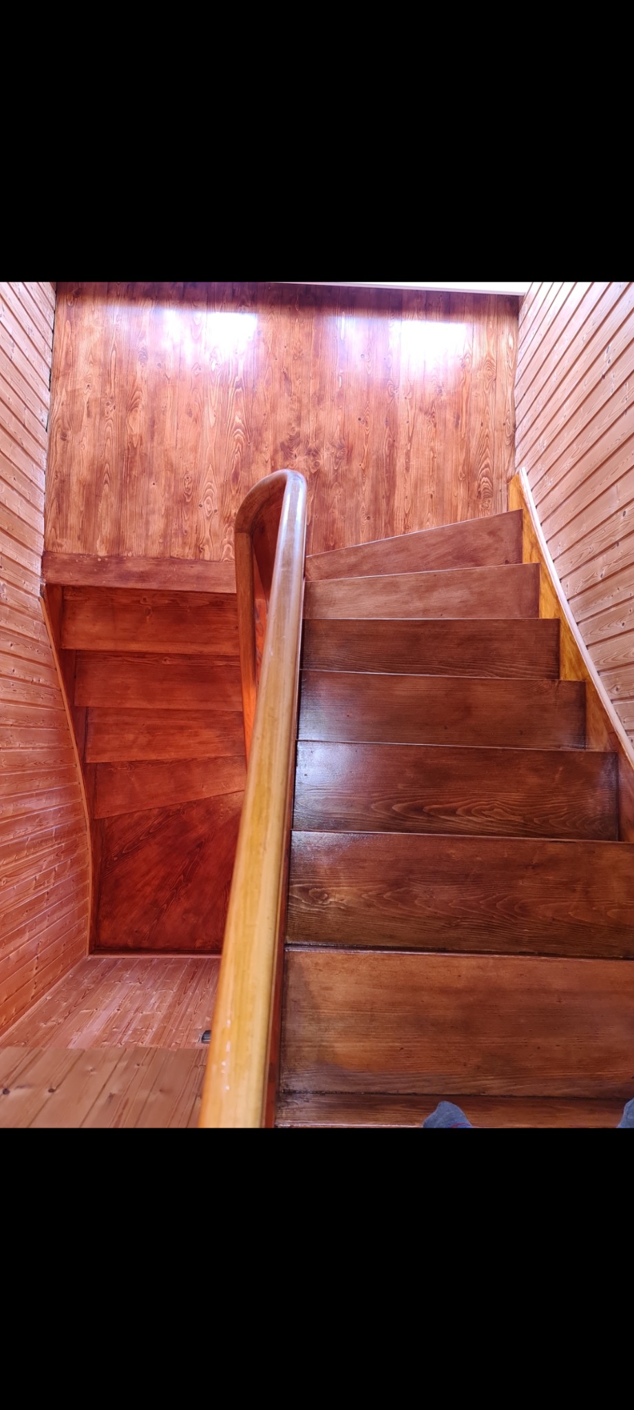 Drewniane schody spiralne z jasną balustradą, wykończone boazerią w odcieniach brązu, widziane z dołu, z fragmentem stopy w skarpetce w dolnej części kadru.