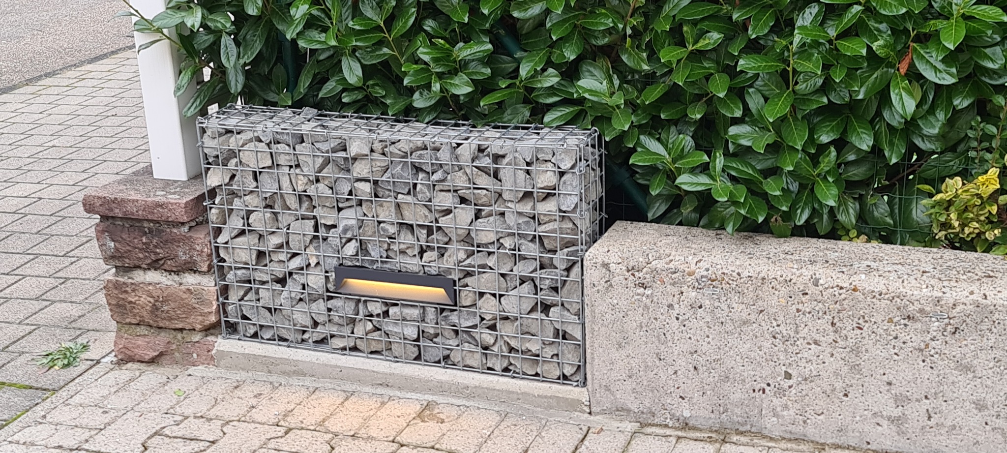 Gabion z oświetleniem LED wkomponowany w ogrodzenie, widok z poziomu chodnika.