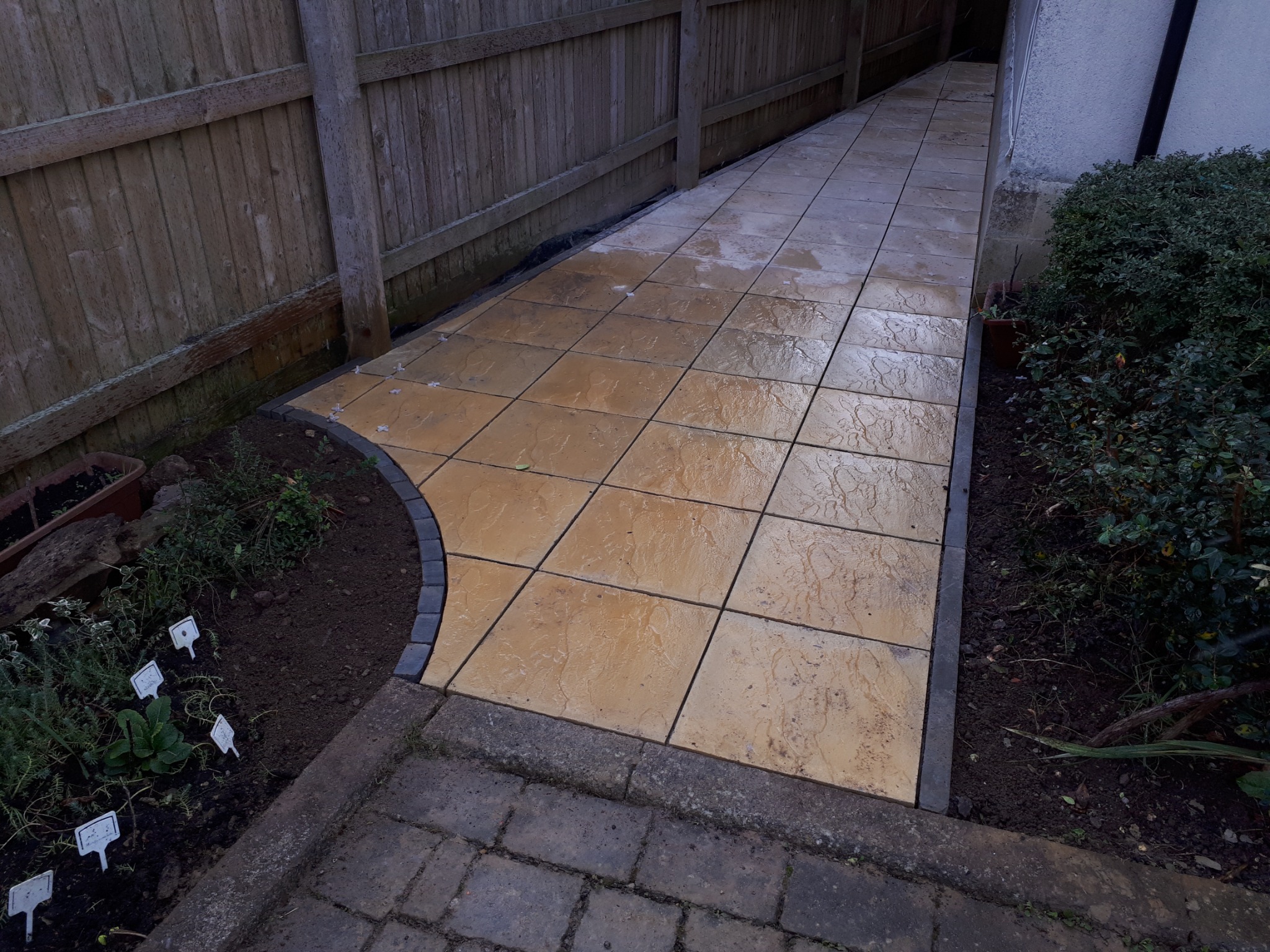 Ułożona ścieżka z kwadratowych, beżowych płyt chodnikowych, obrzeża wykończone ciemnoszarą kostką, widoczne rośliny i drewniany płot w tle.