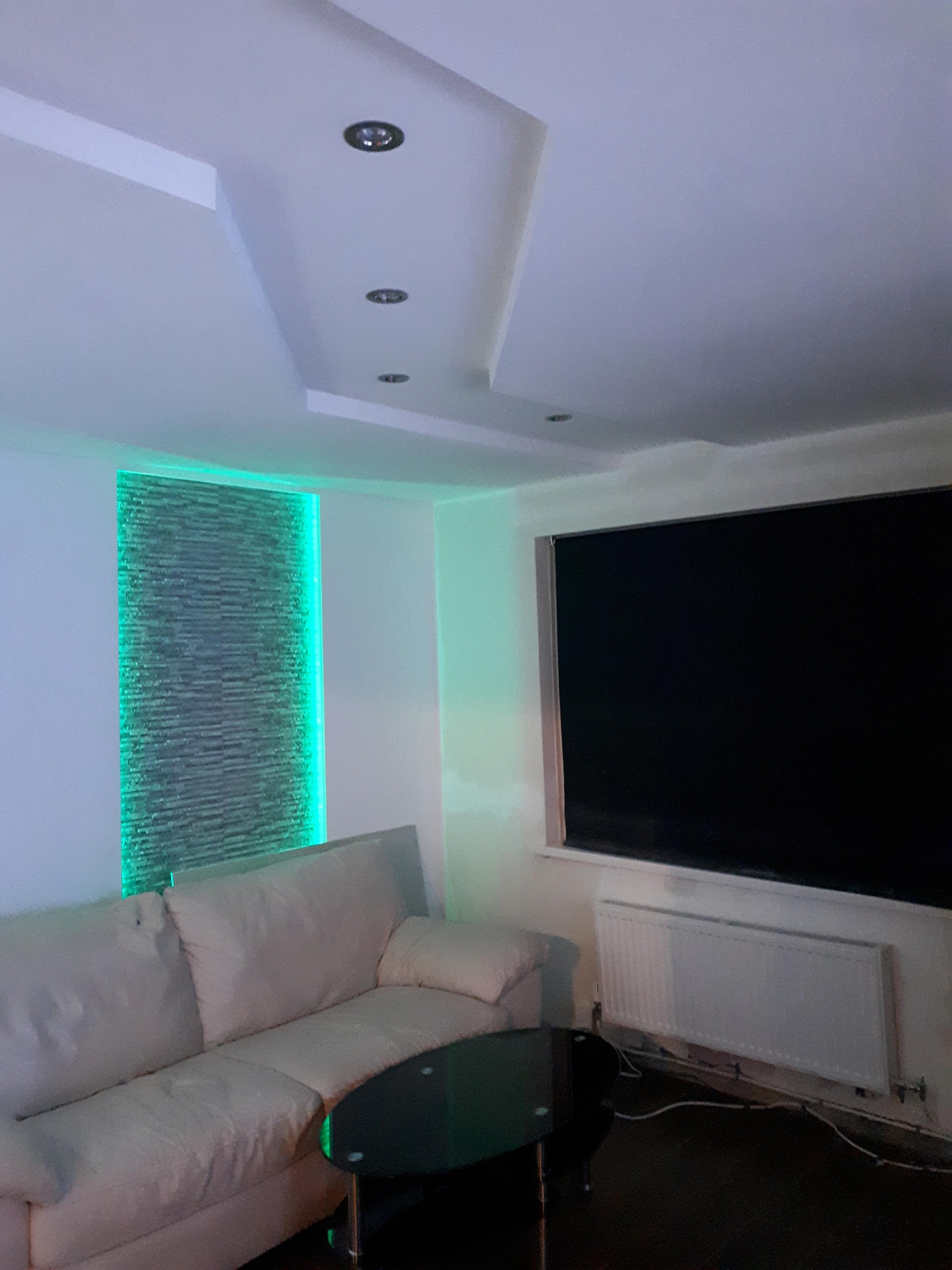 Salon z podświetlaną ścianą z kamienia dekoracyjnego, białym narożnikiem i czarnym stolikiem kawowym. Sufit podwieszany z oświetleniem punktowym.