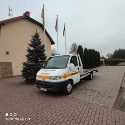Biała laweta z żółtymi oznaczeniami firmy transportowej, zaparkowana na brukowanym placu przed budynkiem z trzema flagami i choinką, pochmurny dzień.