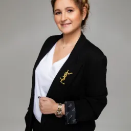 Portret kobiety w czarnym żakiecie z broszką YSL, białym topie i złotym zegarku, z warkoczem na głowie, uśmiechającej się do kamery na szarym tle.