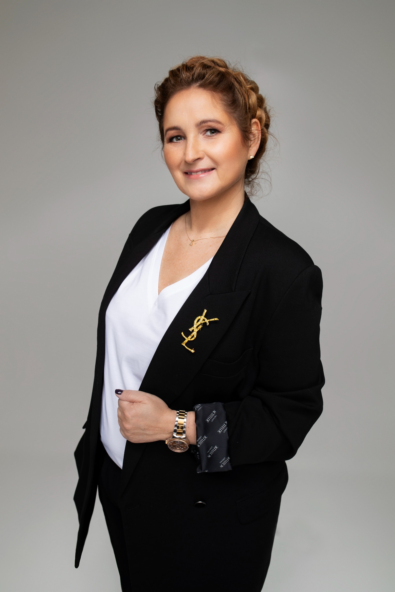 Portret kobiety w czarnym żakiecie z broszką YSL, białym topie i złotym zegarku, z warkoczem na głowie, uśmiechającej się do kamery na szarym tle.