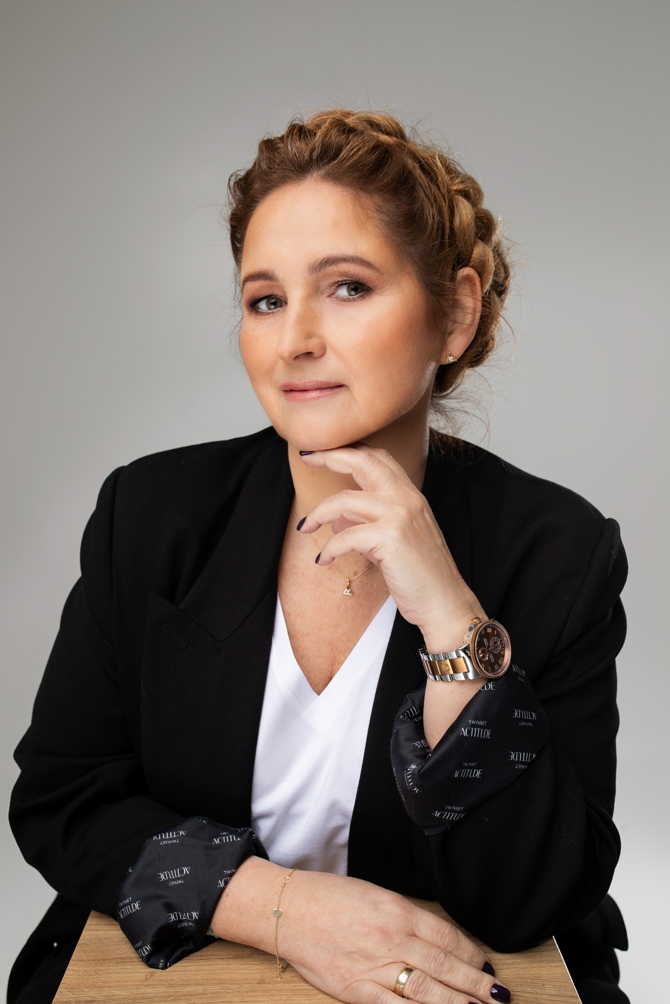 Portret kobiety w czarnym żakiecie, białej koszulce i z upiętymi włosami, z ręką pod brodą i złotym zegarkiem na nadgarstku, na neutralnym tle.