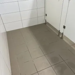 Wnętrze toalety publicznej z szarymi płytkami podłogowymi i białymi ścianami wyłożonymi kafelkami, widoczne kabiny WC z metalowymi wspornikami podtrzymującymi drzwi.