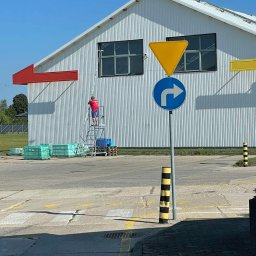 HEFEST GROUP SPÓŁKA Z OGRANICZONĄ ODPOWIEDZIALNOŚCIĄ - Pracownik na aluminiowej drabinie myje okno na wysokości w magazynie z blachy trapezowej, widoczne skrzynki z tworzywa sztucznego i znak drogowy.