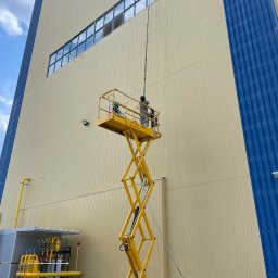 HEFEST GROUP SPÓŁKA Z OGRANICZONĄ ODPOWIEDZIALNOŚCIĄ - Pracownik na żółtym podnośniku koszowym myje okna na wysokości w przemysłowym budynku o beżowo-niebieskiej elewacji. Widoczny długi kij teleskopowy z myjką do okien.