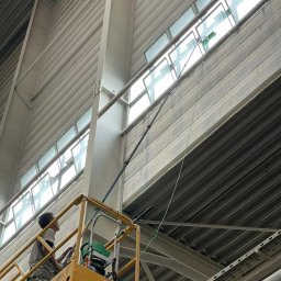 HEFEST GROUP SPÓŁKA Z OGRANICZONĄ ODPOWIEDZIALNOŚCIĄ - Pracownik na podnośniku koszowym myje wysoko położone okna w hali przemysłowej za pomocą długiej lancy z zieloną końcówką, widoczne smugi brudu na oknach przed umyciem.