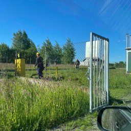 HEFEST GROUP SPÓŁKA Z OGRANICZONĄ ODPOWIEDZIALNOŚCIĄ - Dwóch pracowników w kaskach przycinających trawę wokół ogrodzenia terenu przemysłowego w słoneczny dzień.