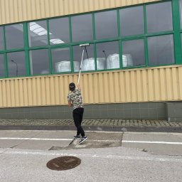 HEFEST GROUP SPÓŁKA Z OGRANICZONĄ ODPOWIEDZIALNOŚCIĄ - Mężczyzna w koszulce w kwiaty myje okna na wysokości w budynku przemysłowym za pomocą długiej myjki, widoczne worki wewnątrz budynku.