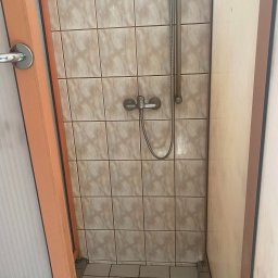 HEFEST GROUP SPÓŁKA Z OGRANICZONĄ ODPOWIEDZIALNOŚCIĄ - Wnętrze kabiny prysznicowej z beżowymi płytkami, widoczna bateria i słuchawka prysznicowa, fragment drzwi z pomarańczową framugą po lewej stronie.