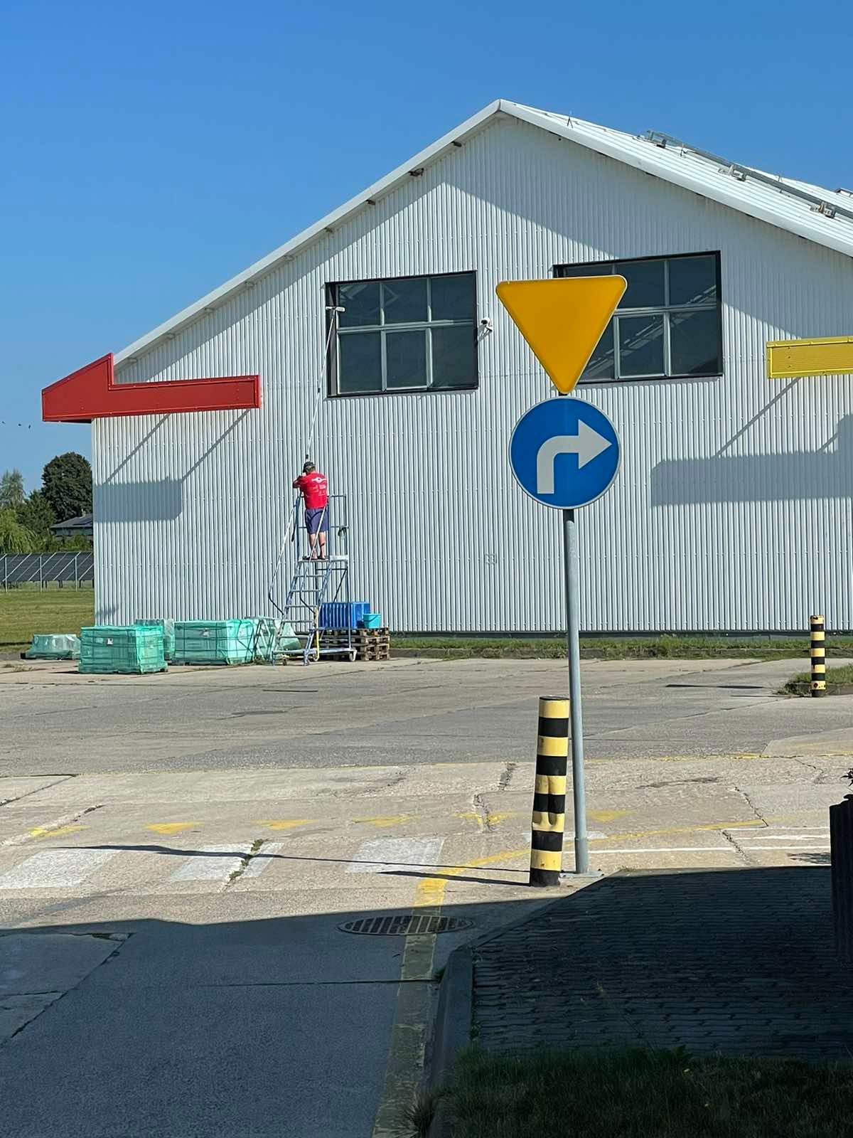 Pracownik na aluminiowej drabinie myje okno na wysokości w magazynie z blachy trapezowej, widoczne skrzynki z tworzywa sztucznego i znak drogowy.