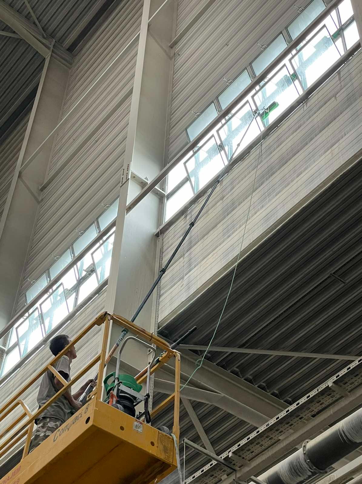 Pracownik na podnośniku koszowym myje wysoko położone okna w hali przemysłowej za pomocą długiej lancy z zieloną końcówką, widoczne smugi brudu na oknach przed umyciem.