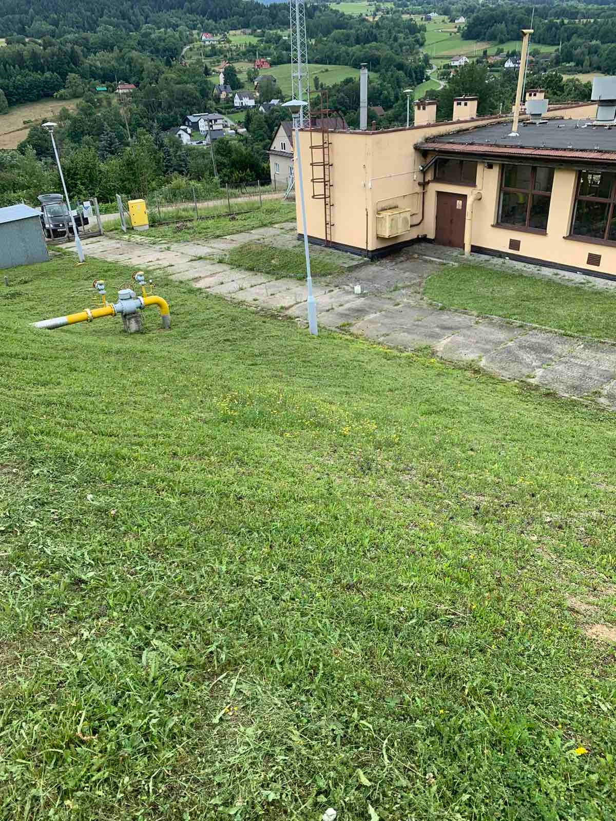 Nierównomiernie skoszona trawa przed budynkiem z żółtą instalacją gazową i widokiem na zalesione wzgórza w tle.