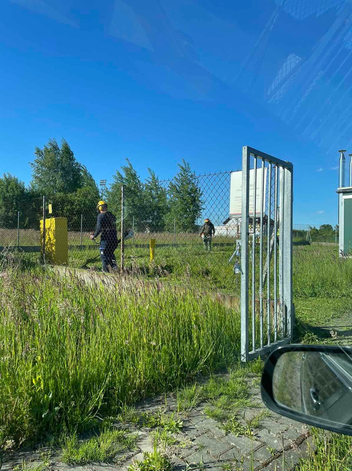 Dwóch pracowników w kaskach przycinających trawę wokół ogrodzenia terenu przemysłowego w słoneczny dzień.