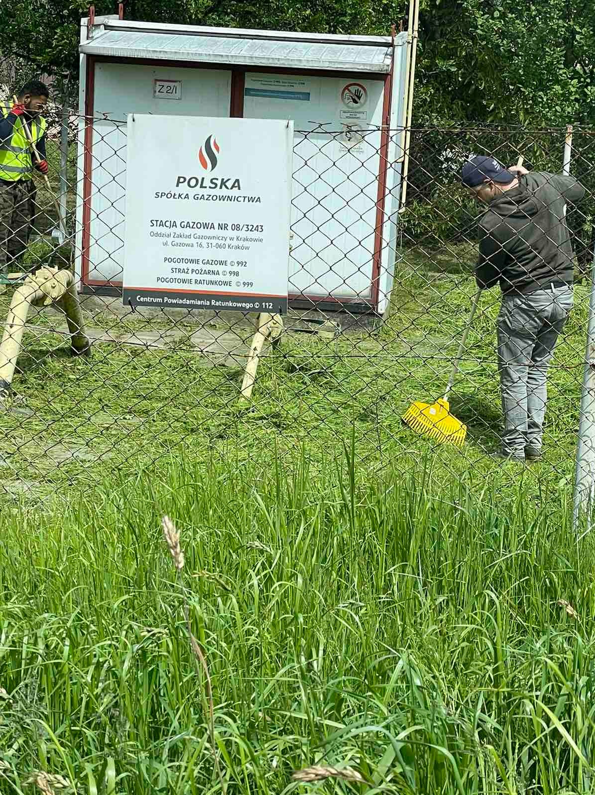 Pracownik w czapce grabi trawę przy ogrodzeniu stacji gazowej z tablicą informacyjną, w tle drugi pracownik i budynek stacji.