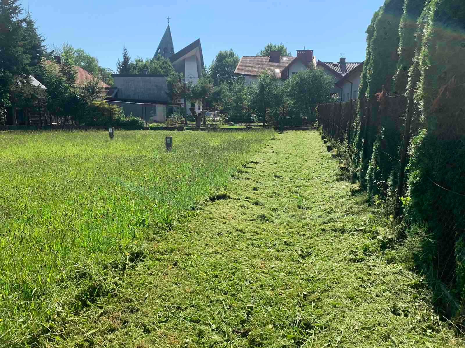 Połowa trawnika skoszona, z wyraźną linią podziału między wysoką a krótko przystrzyżoną trawą, w tle zabudowa wiejska z kościołem.