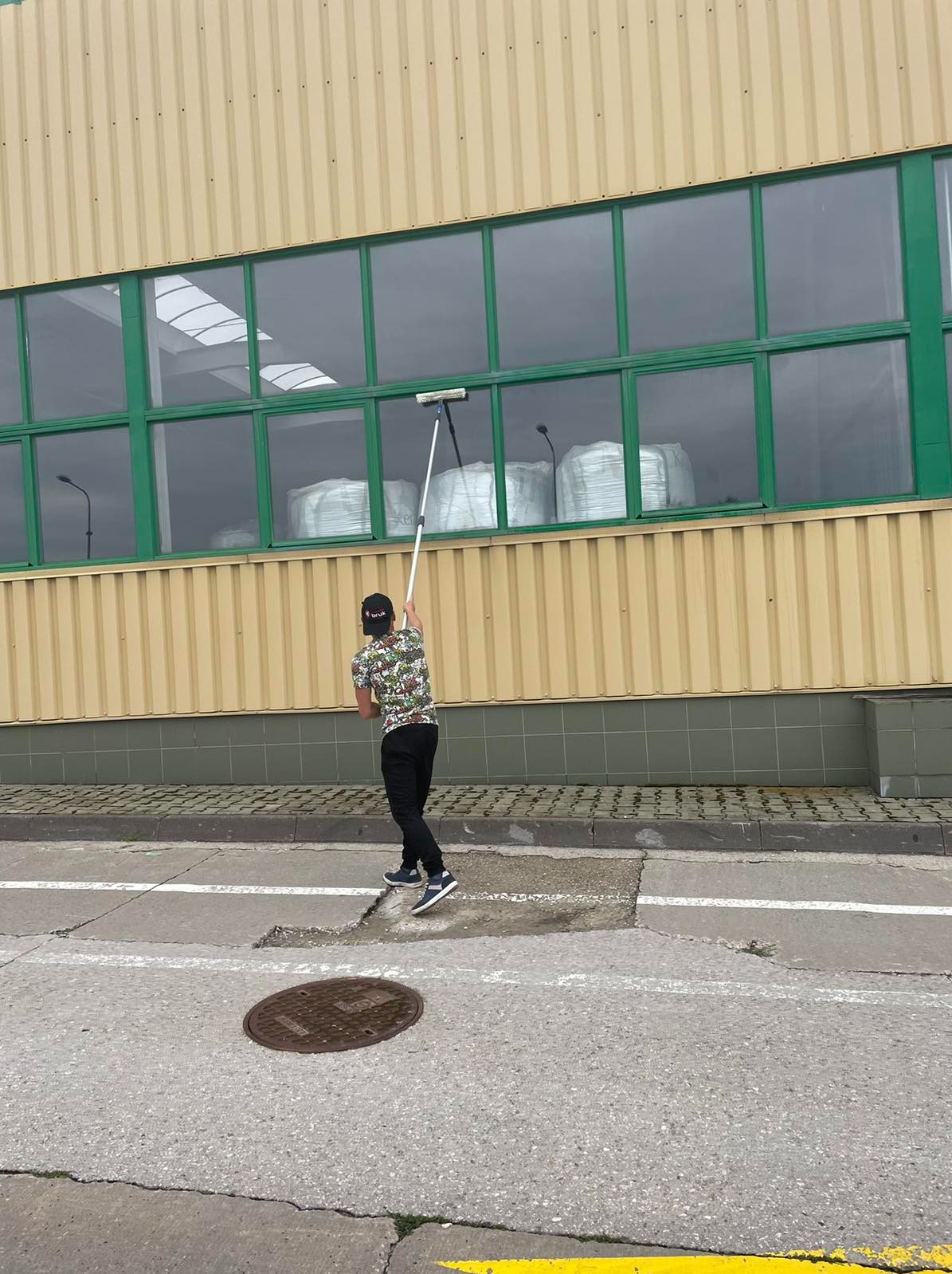 Mężczyzna w koszulce w kwiaty myje okna na wysokości w budynku przemysłowym za pomocą długiej myjki, widoczne worki wewnątrz budynku.