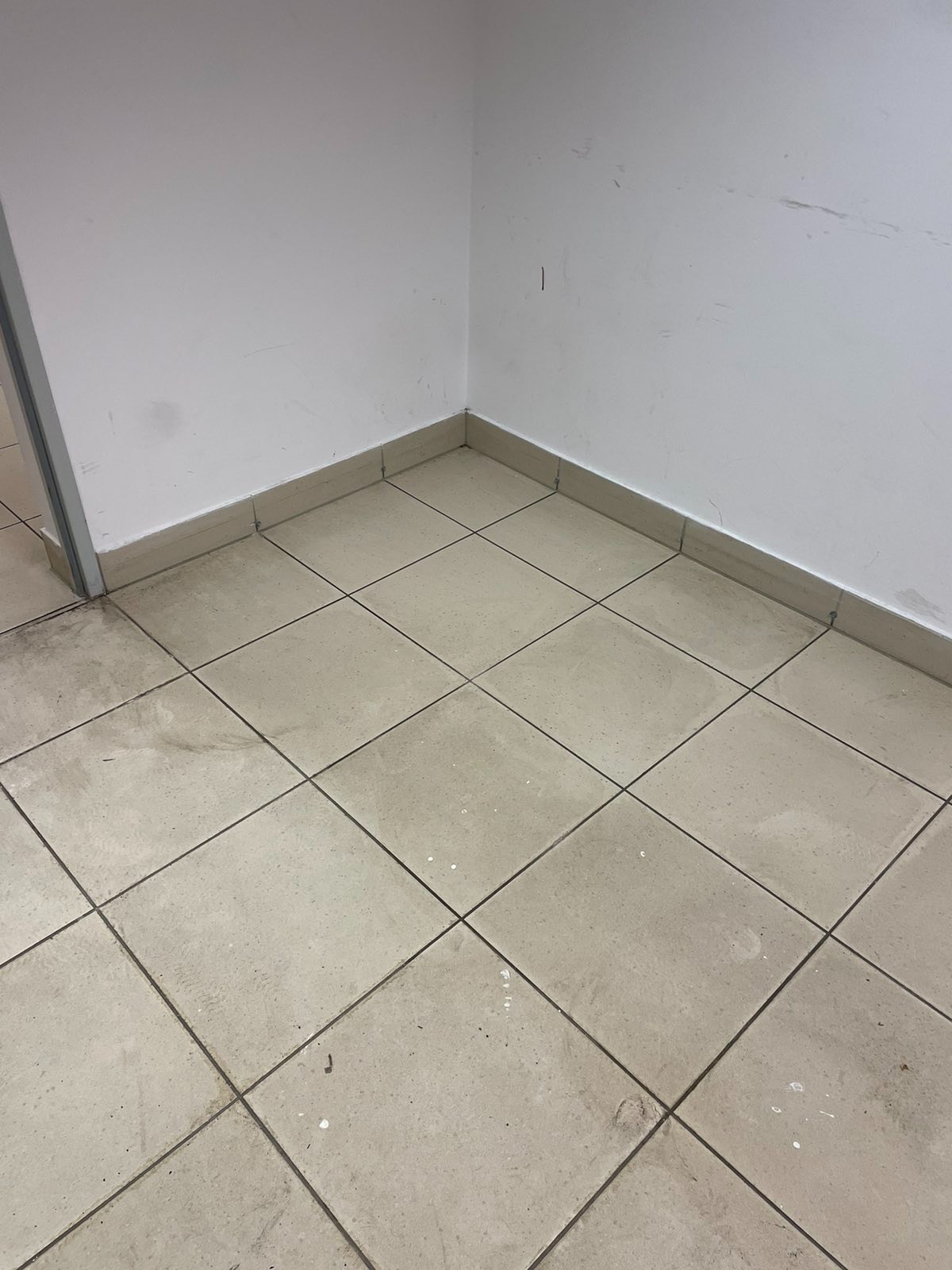 Fragment rogu pomieszczenia z beżowymi, kwadratowymi płytkami podłogowymi z widocznymi zabrudzeniami i białymi ścianami z drobnymi śladami.