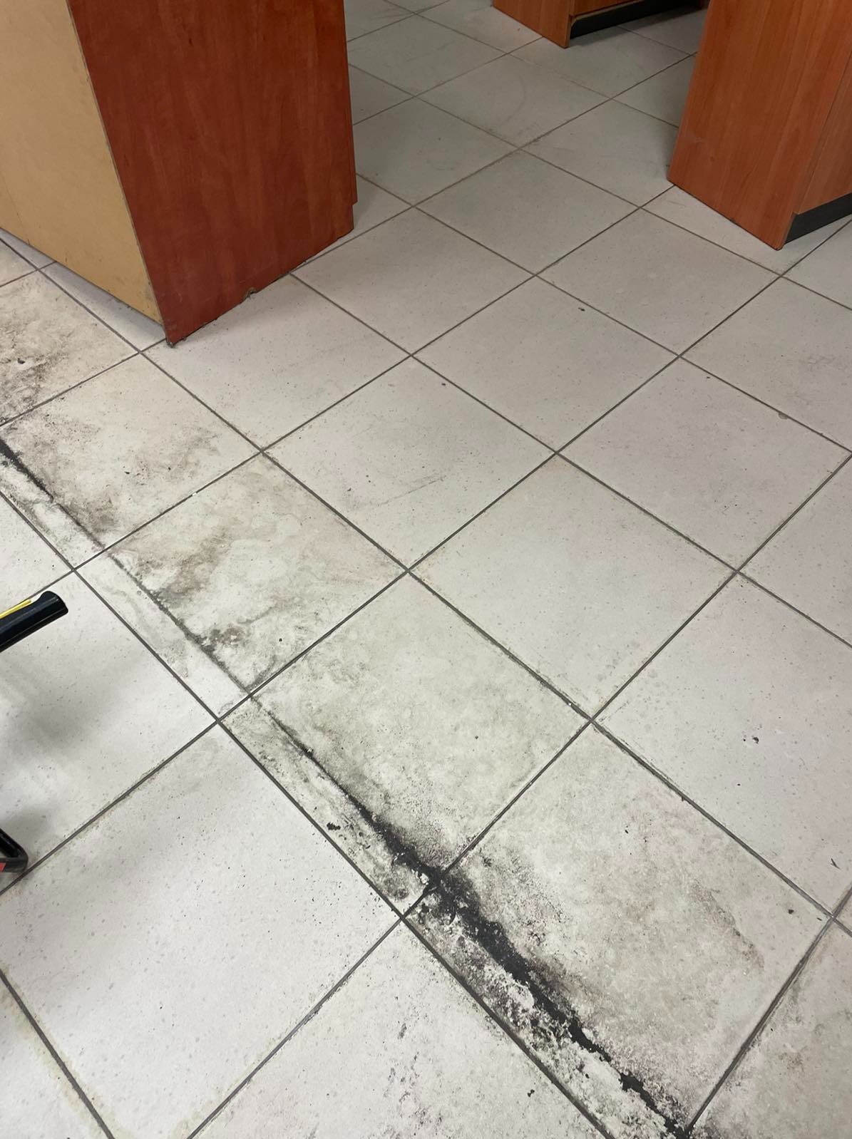 Silnie zabrudzona podłoga z płytek ceramicznych w biurze, widoczne ciemne smugi i plamy wzdłuż fug, częściowo oczyszczona w lewym dolnym rogu.